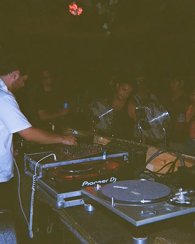 Un DJ en t-shirt blanc aux platines du Chapiteau, photographié à l'appareil jetable