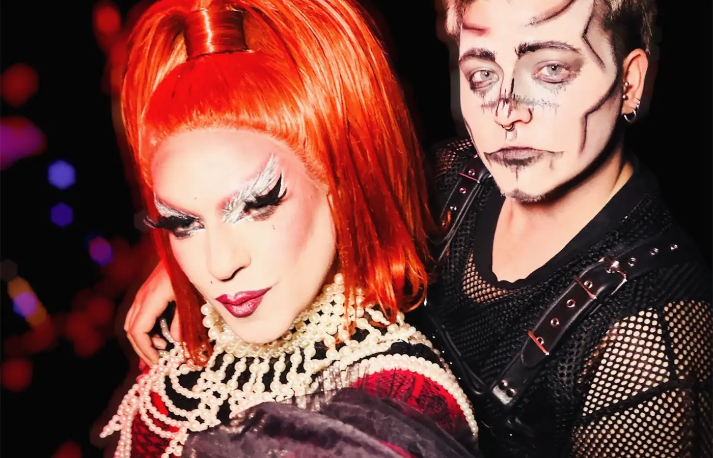Une drag queen en perruque rouge et colliers de perles pose avec un artiste maquillé en squelette, sous des confettis, à Speechless