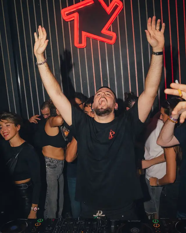 Un DJ bras levés derrière les platines du Baby Club, sous le logo rouge en néon