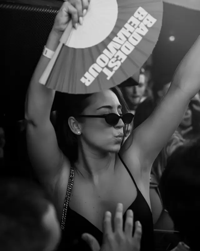 En noir et blanc, une jeune femme en lunettes de soleil brandit un éventail « Baddest Behaviour » au Barta